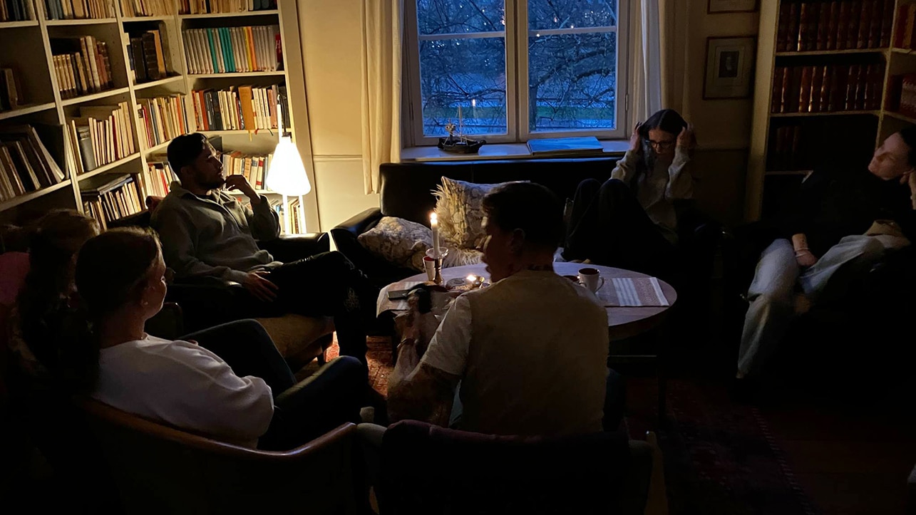 Några personer som sitter runt ett bord i soffor. Utanför fönstret är det skymning. Det ser mysigt ut.